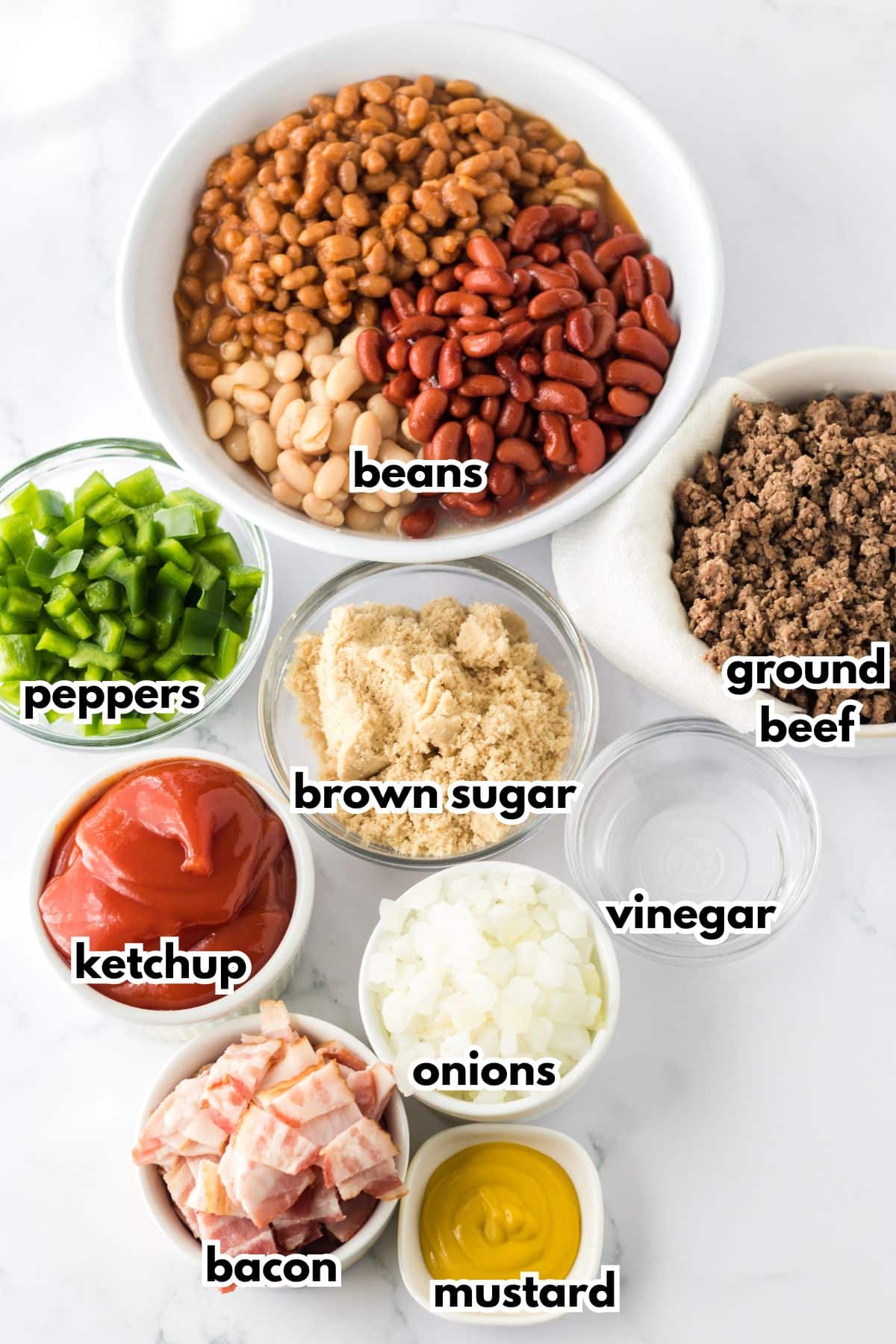 bowls of beans, peppers, brown sugar, ketchup, bacon, onions, mustard, vinegar, and ground beef on a counter.