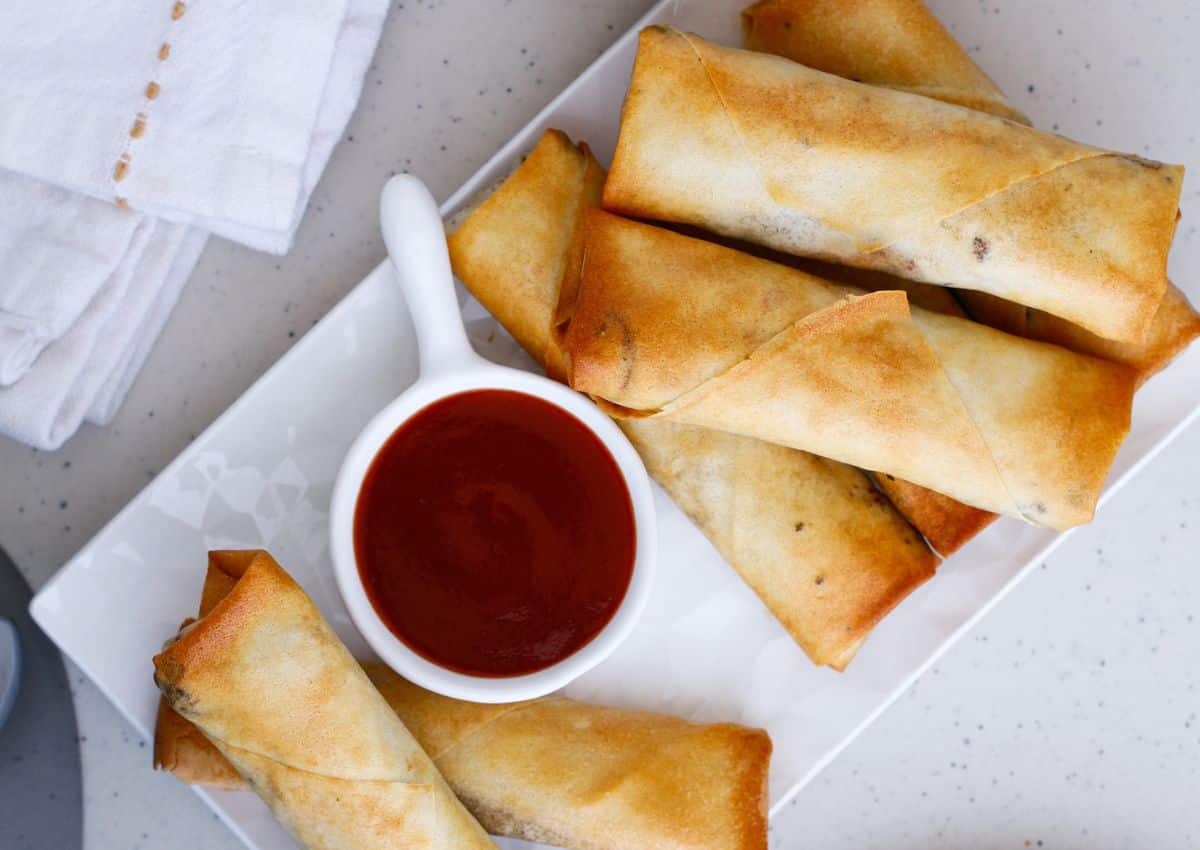 Egg rolls stacked on a serving patter with some sweet and sour sauce for dipping.