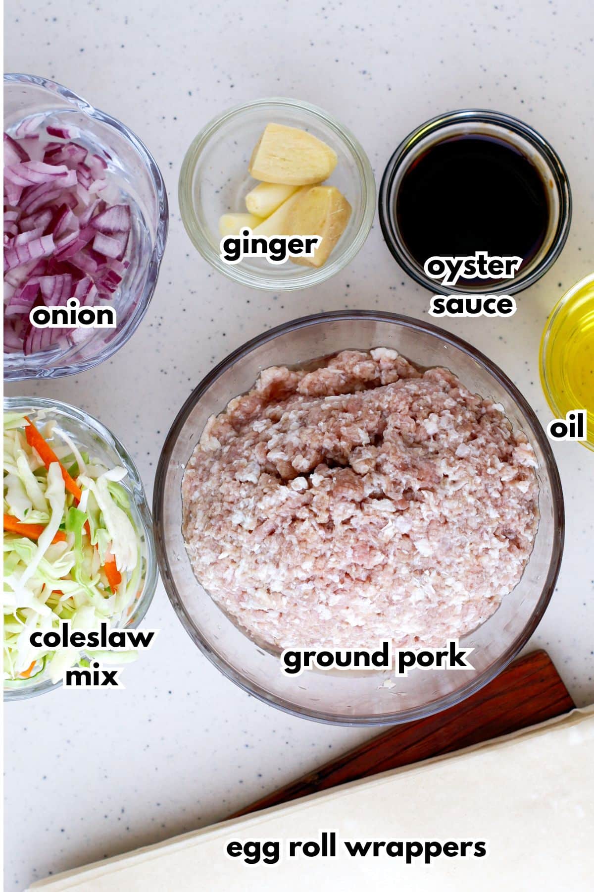Bowls of onion, colelslaw mix,ginger, oyster sauce, ground pork, oil, and egg roll wrappers on a counter.