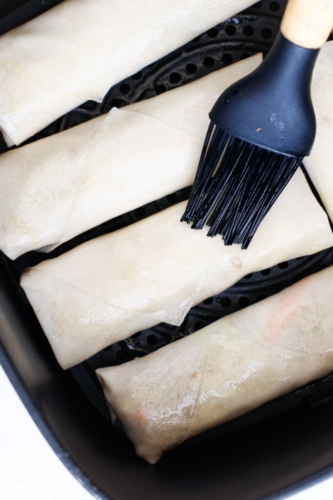 oil being brushed on top of egg rolls in the air fryer.