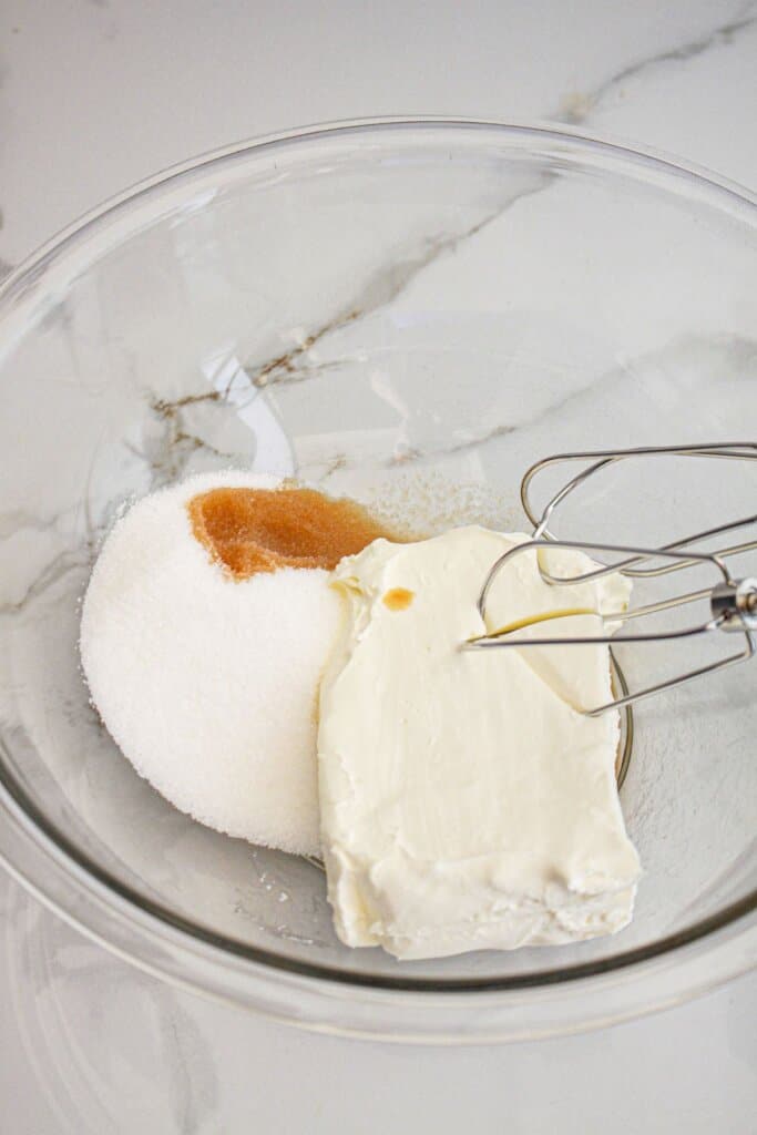 Cream cheese, sugar, and vanilla being mixed in a glass mixing bowl.