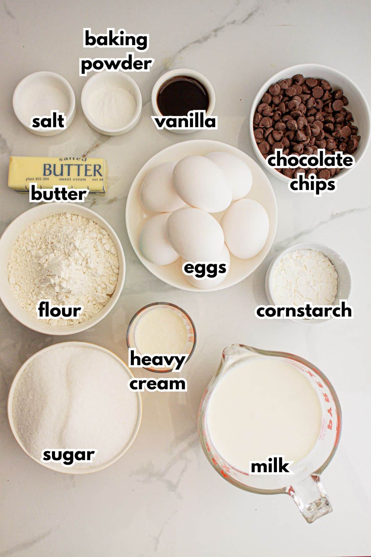 Bowls of salt, baking powder, vanilla, butter, chocolate chips, flour, heavy cream, sugar, milk, and constarch on a counter.