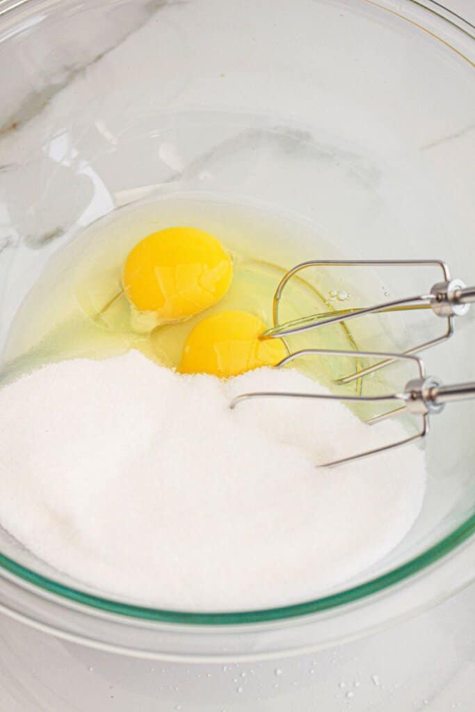 Eggs and sugar being mixed together with a hand mixer in a large glass mixing bowl.