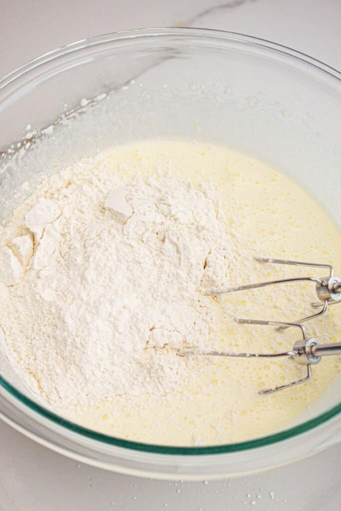 Dry ingredients being added to batter with a hand mixer in a glass mixing bowl.