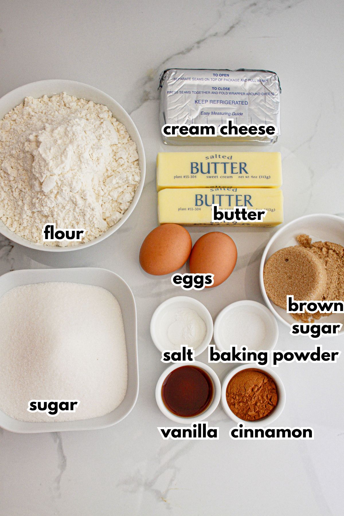 Bowls of flour, sugar, cream cheese, butter, eggs, salt, baking powder, vanilla, cinnamon and brown sugar on a counter.