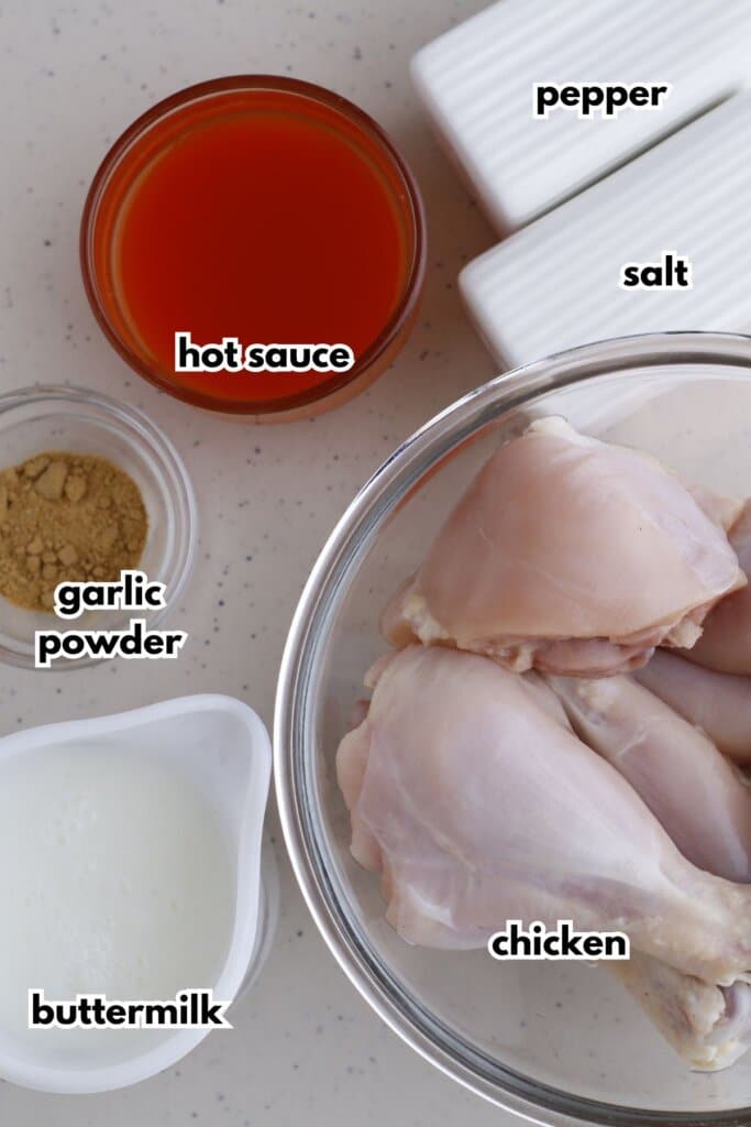 Bowls of salt, pepper, hot sauce, garlic powder, buttermilk, and chicken pieces on a counter.