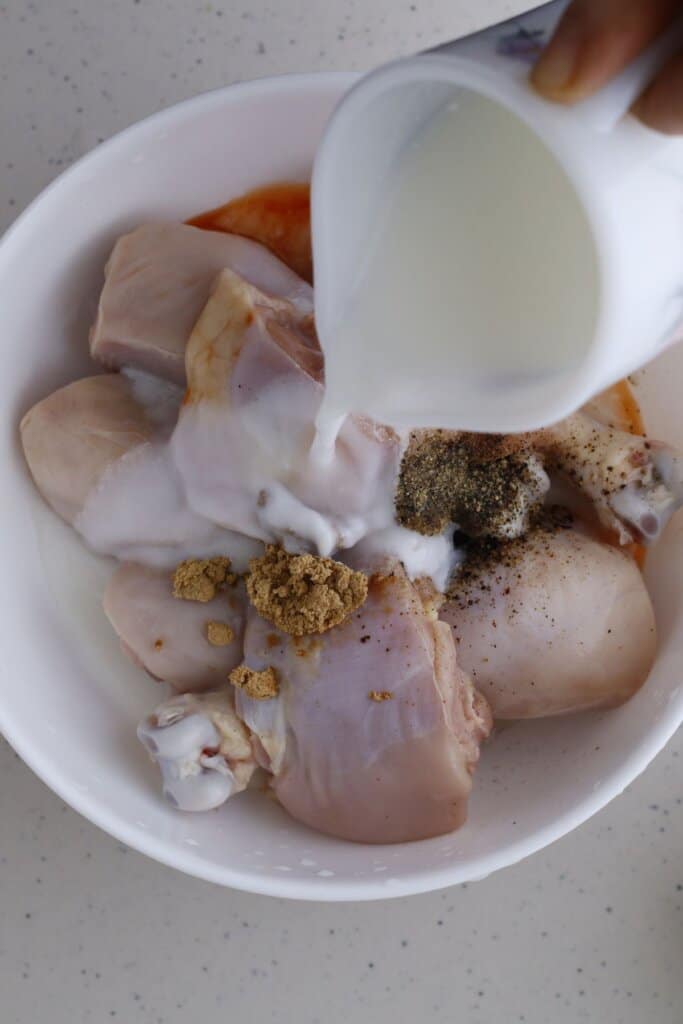 Buttermilk being poured over seasoning chicken pieces in a mixing bowl.