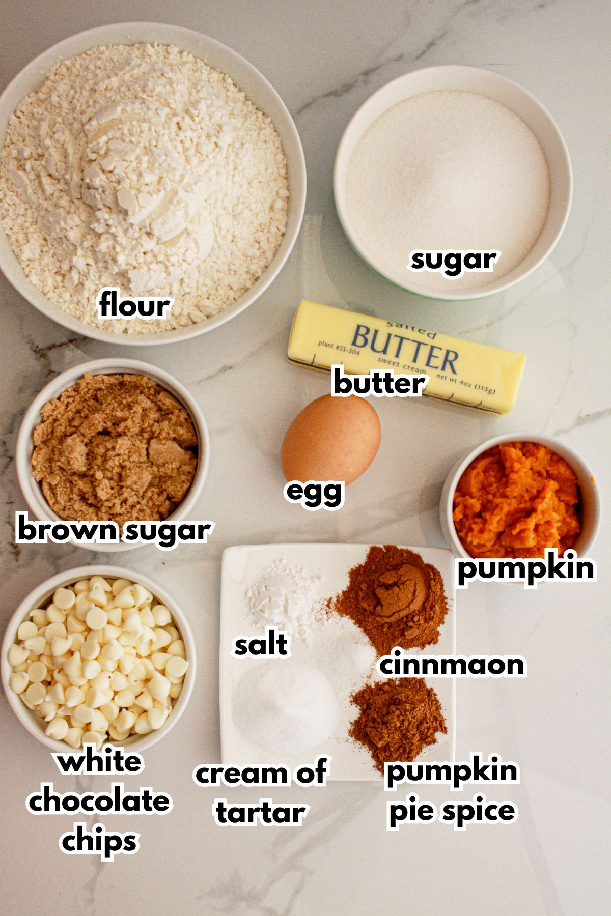 Bowls of flour, sugar, brown sugar, egg, butter, white chocolate chip, salt, cream of tarter, pumpkin, cinnamon and pumpkin pie spice.