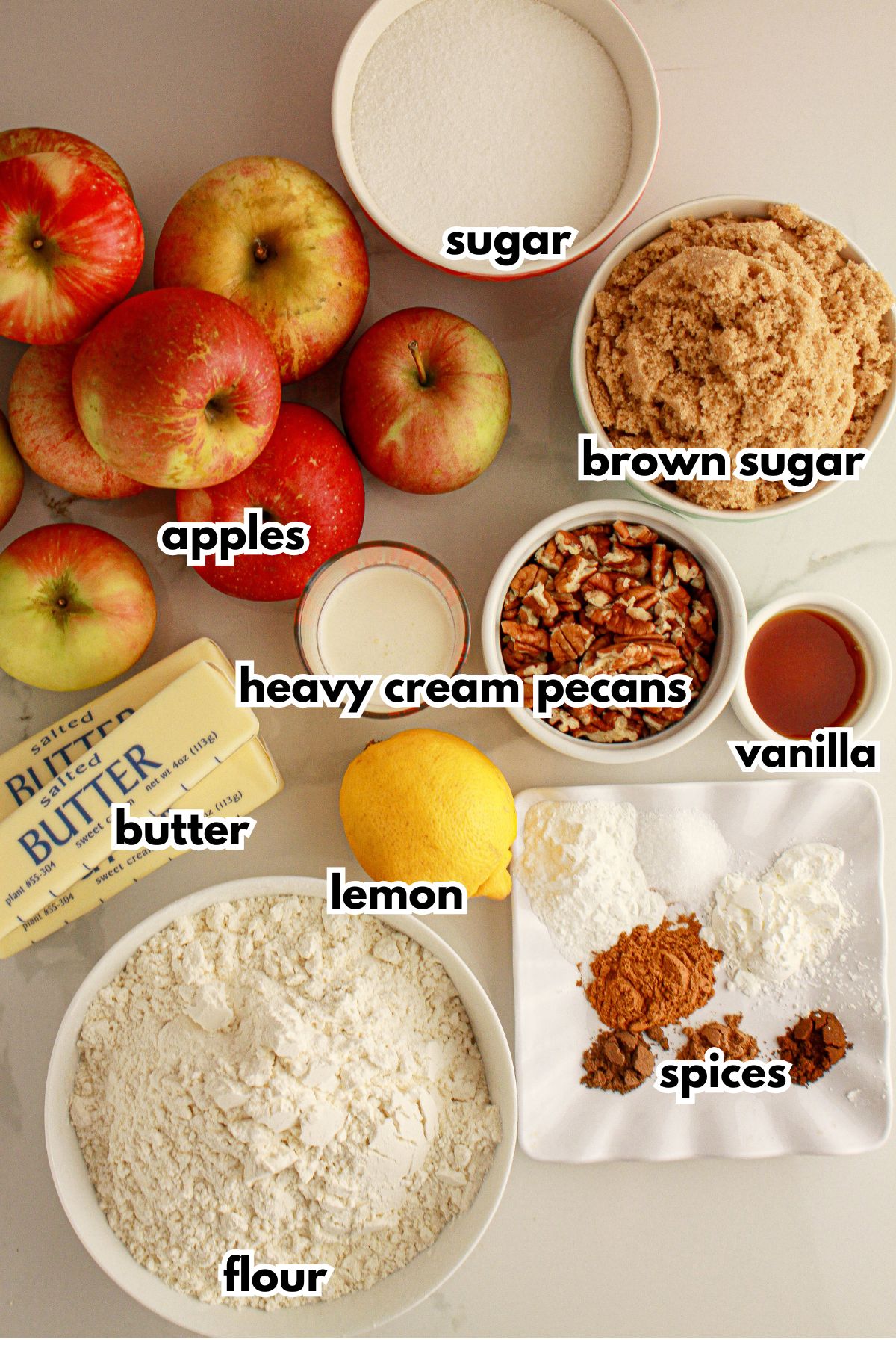 Apples, and bowls of sugar, brown sugar, butter, heavy cream, pecans, vanilla, lemon, flour, spices and vanilla extract on a counter.