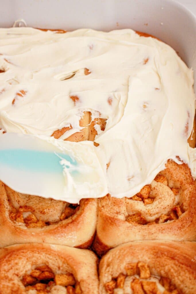 Cream cheese frosting being spread on top of cinnamon rolls in a baking dish.