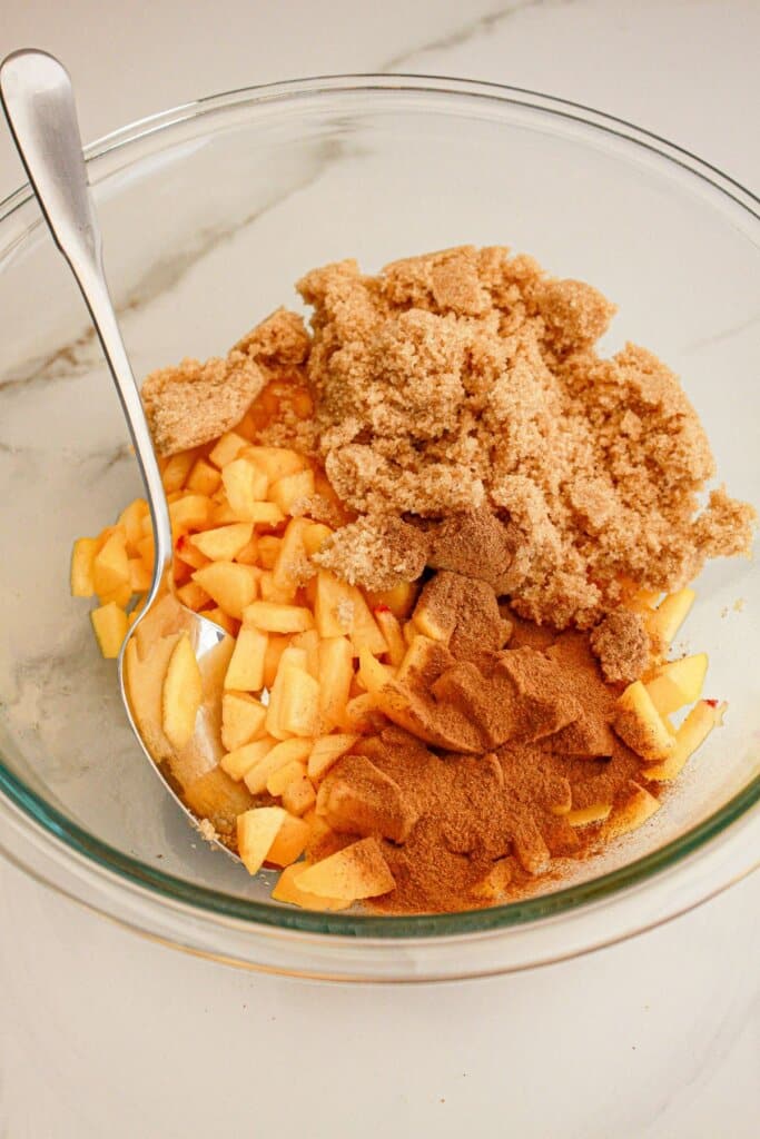 Brown sugar, peaches, cinnamon and nutmeg being mixed in a glass mixing bowl.
