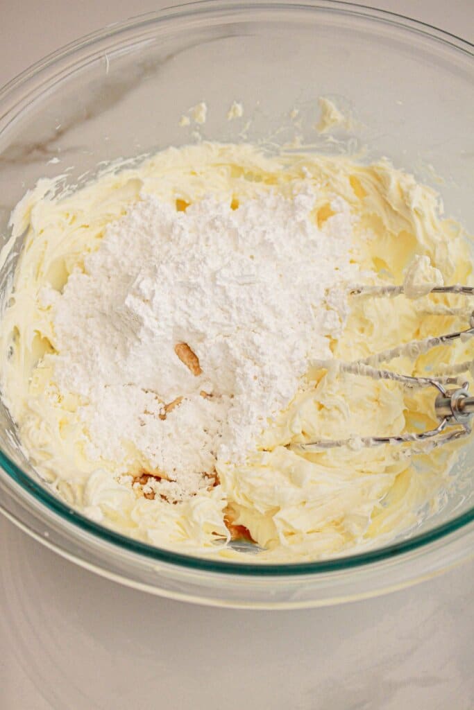 Powdered sugar being added to cream cheee frosting in a glass mixing bowl.
