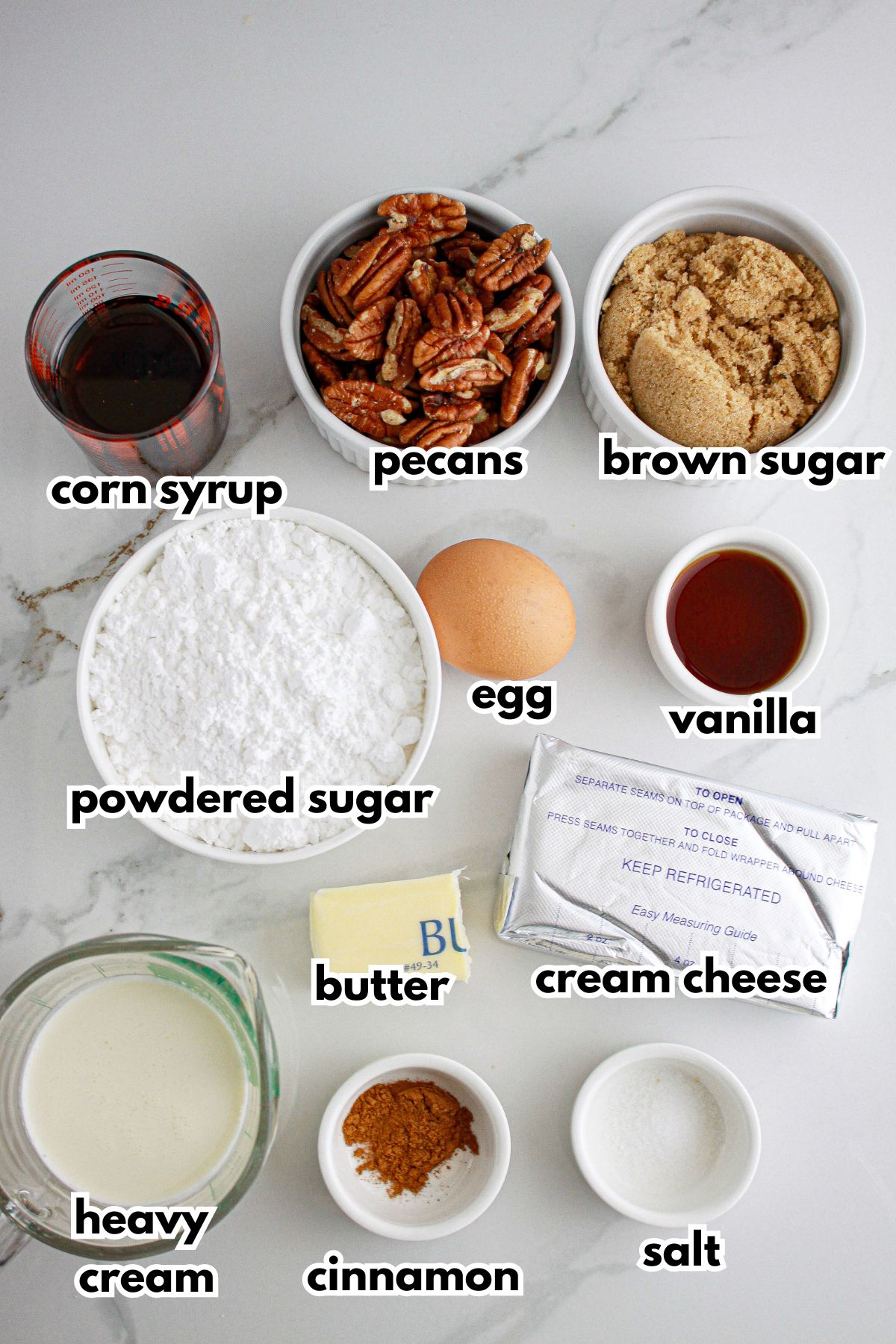 Bowls of corn syrup, pecans, brown sugar, powdered sugar, egg, vanilla, butter, cream cheese, heavy cream, cinnamon, and salt on a counter.