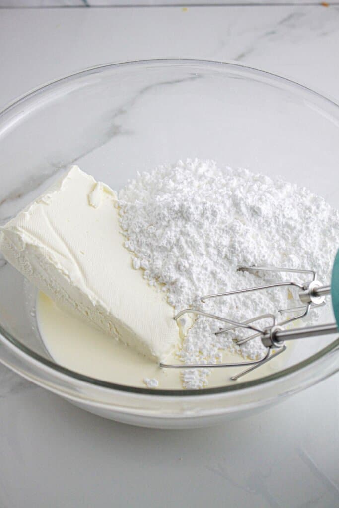 Cream cheese, heavy cream, and powdered sugar being mxied together in a large glass bowl.