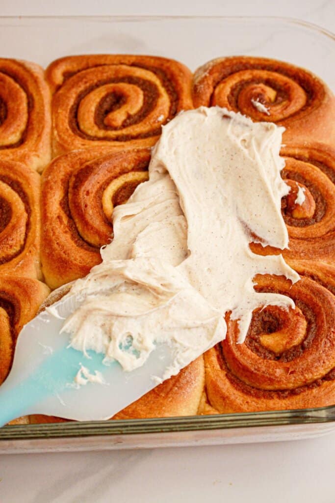Cream cheese frosting being spread on top of cinnamon rolls in a baking dish.