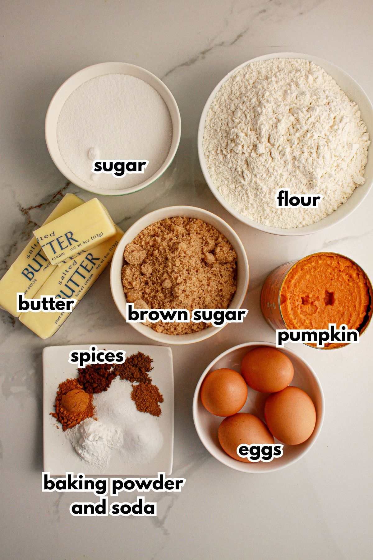 Bowls of sugar, flour, butter, brown sugar, pumpkin, spices, baking powder and soda and eggs on a counter.