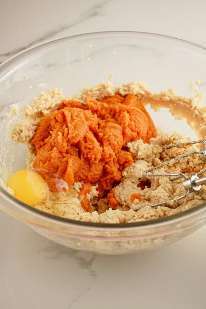 Eggs, and pumpkin being added to butter and sugar in a glass mixing bowl.
