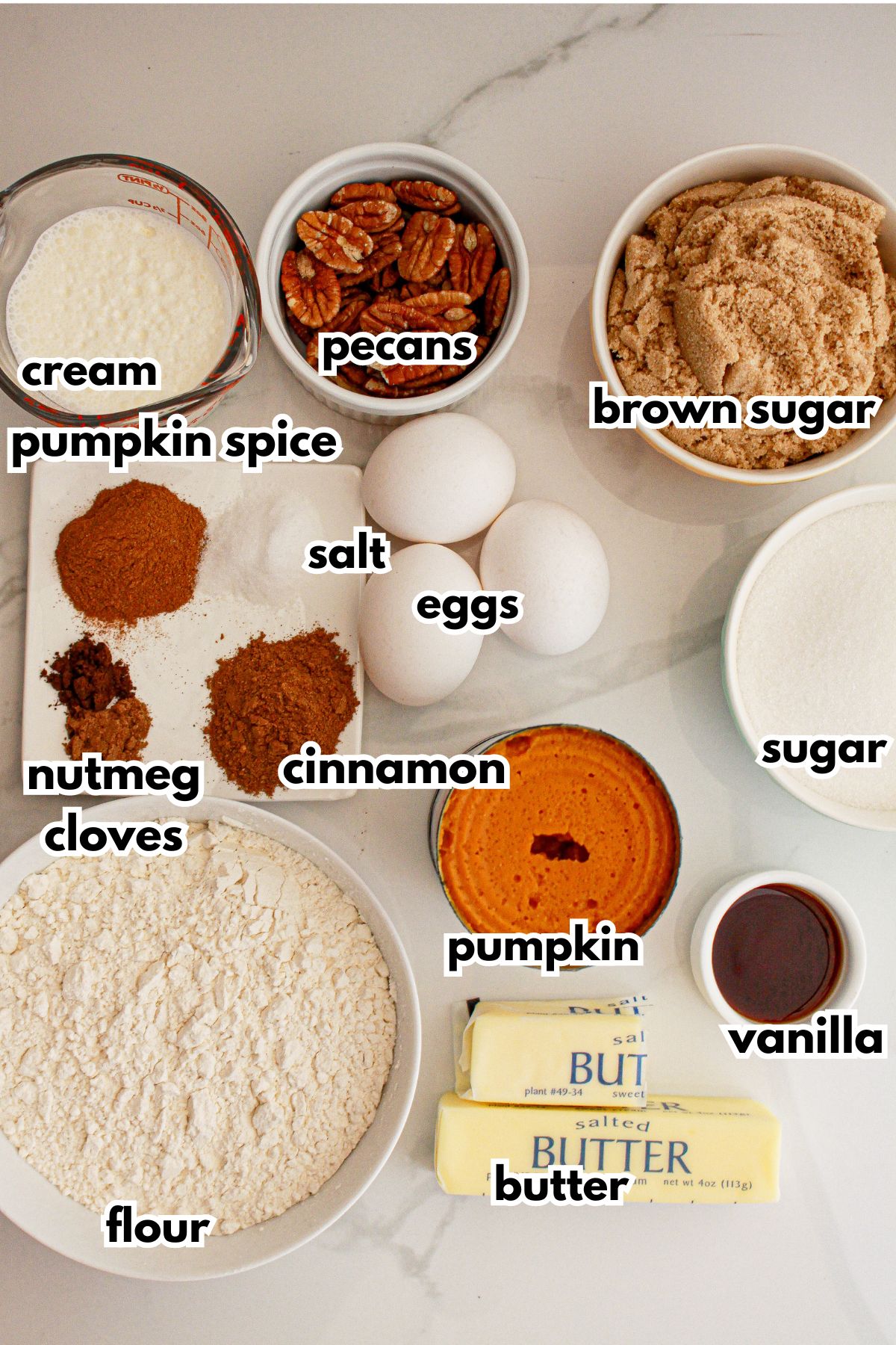 Bowls of cream, pecans, brown sugar, pumpkin spice, salt, nutmeg, cloves, cinnamon, pumpkin puree, sugar, vanilla, butter, and flour on a counter.