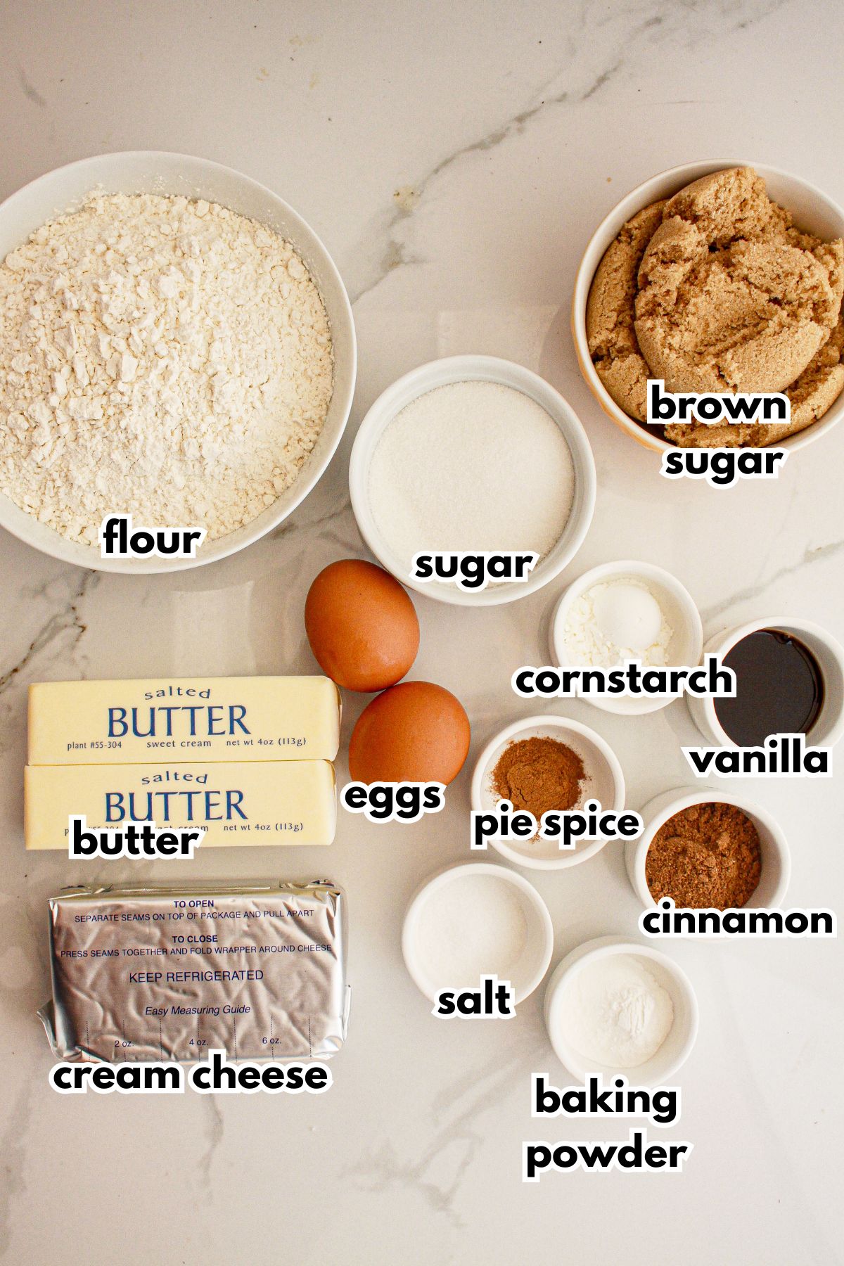 Bowls of flour, sugar, brown sugar, butter, eggs, cornstarch, vanilla, eggs, pie spice, cinnamon, salt, and baking powder on a counter.