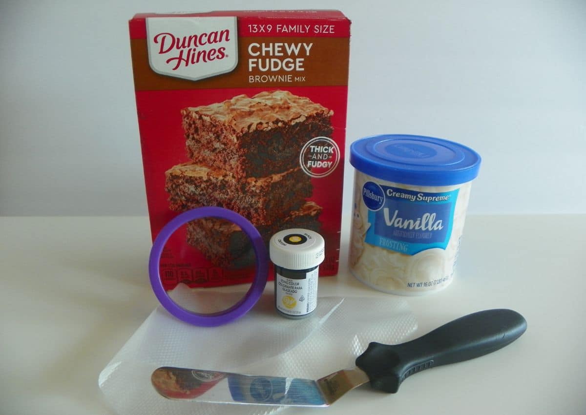 Box of brownie mix, can of vanilla frosting, food coloring, a cookie cutter and an icing spread on a counter.