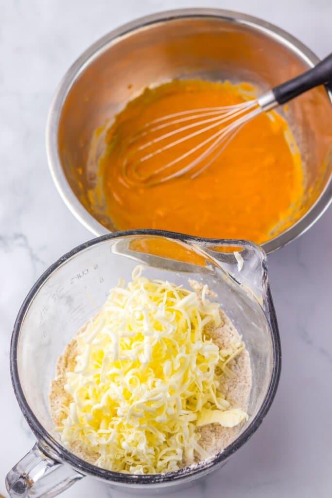 Grated butter being added to dry ingredients in a glass mearsuring cup.