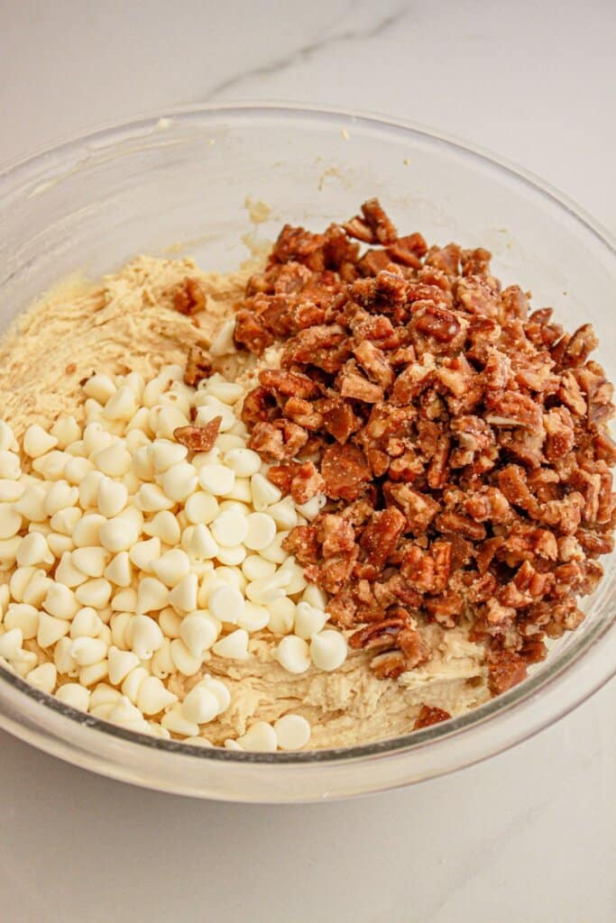 Pecans and white chocolate chips being added to dough in a glass bowl.