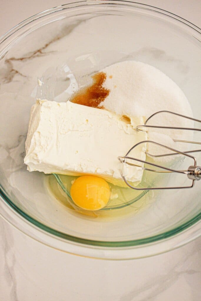 Cheese, sugar, egg, and vanilla being mixed in a glass bowl.