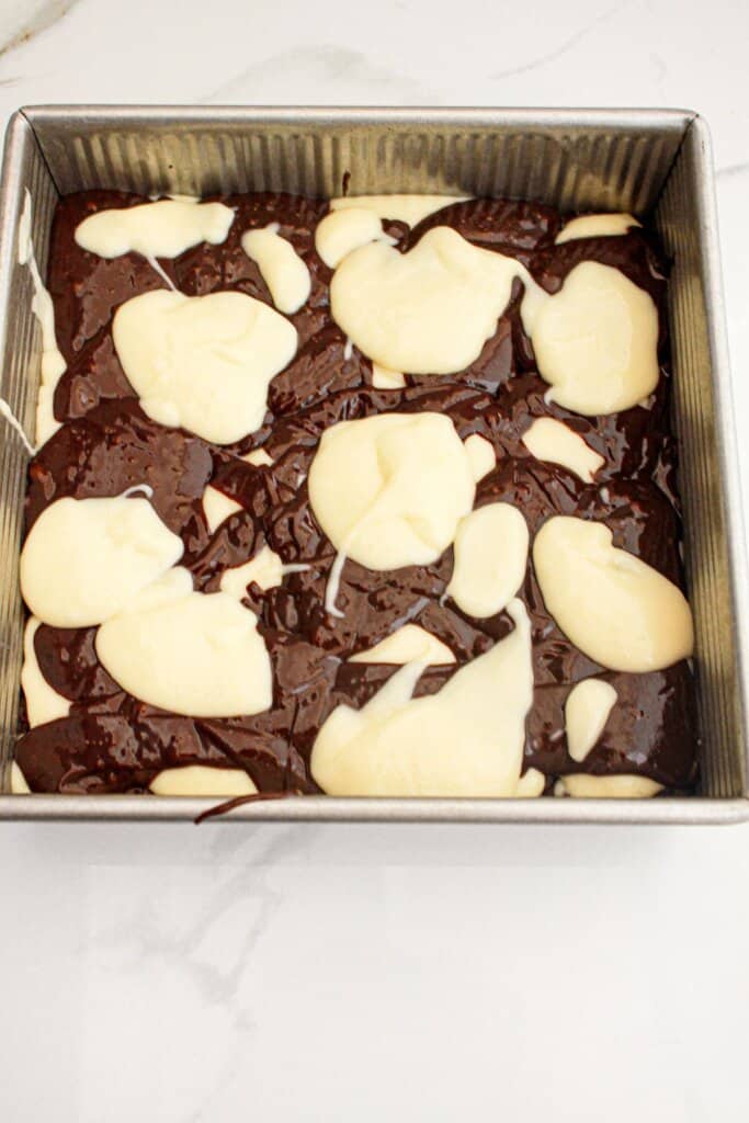 Brownie batter and cream cheese being layered in a baking pan.