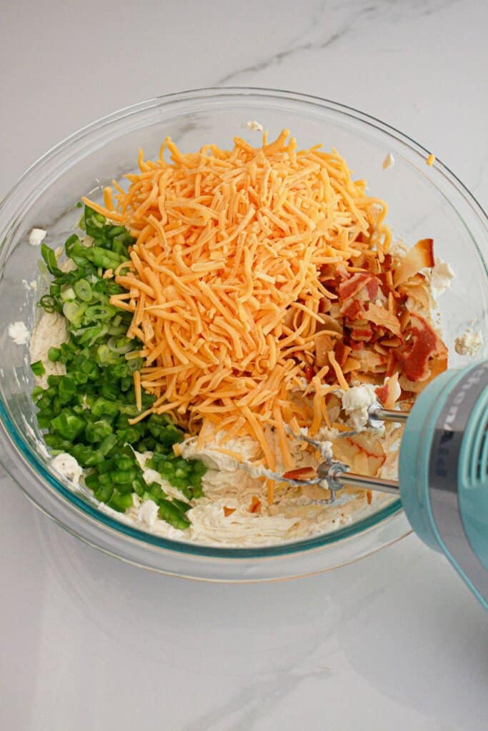 Cheese, bacon, green onions, and cream cheese being mixed in a glass bowl.