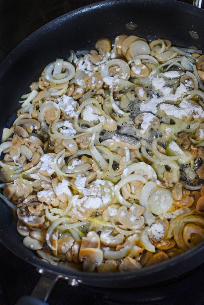 Flour being added on top of vegetables in a skillet.