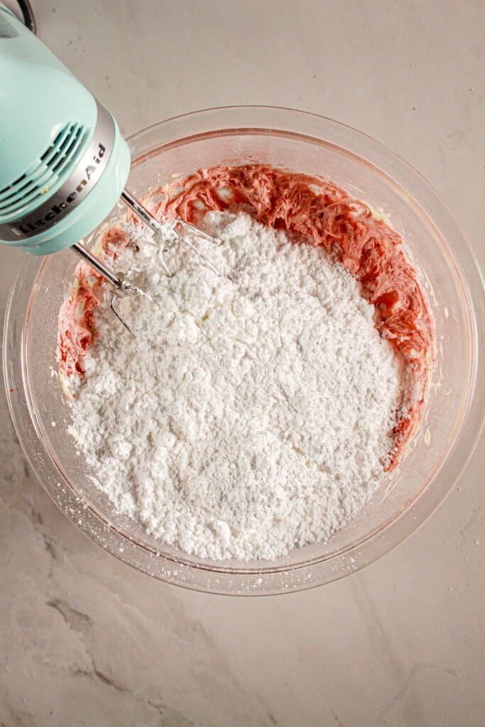 Powdered sugar being added to frosting in a mixing bowl.