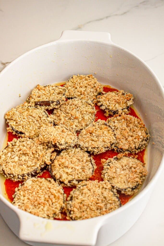 Breaded eggplant slices in a baking dish on top of sauce.