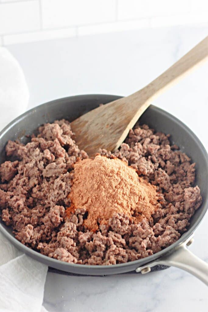 Taco seasoning being added to cooked meat in a skillet.