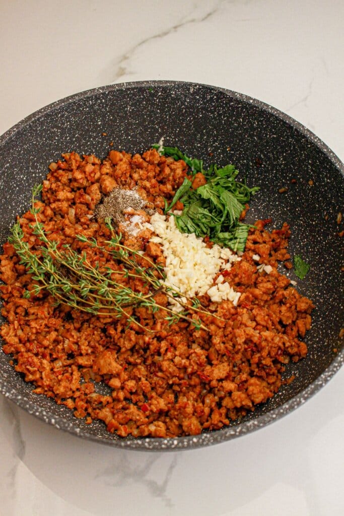 Cooked and crumbled meat, thyme, garlic, and basil in a skillet.