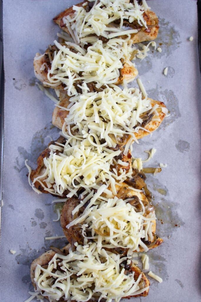 Cheese being sprinkled on top of chicken on a parchment lined baking sheet.