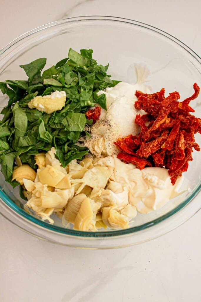 Spinach, sundried tomatoes, and artichoke hearts in a large glass bowl.