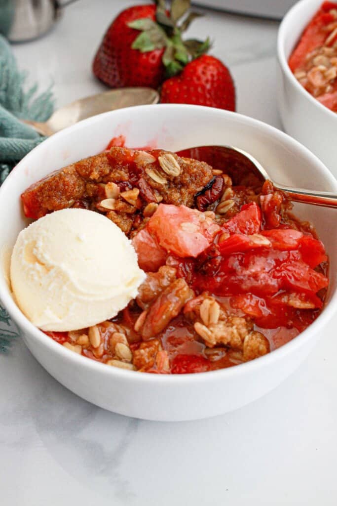 Serving of strawberry rhubarb crisp in a white bowl topped with a scoop of ice cream.