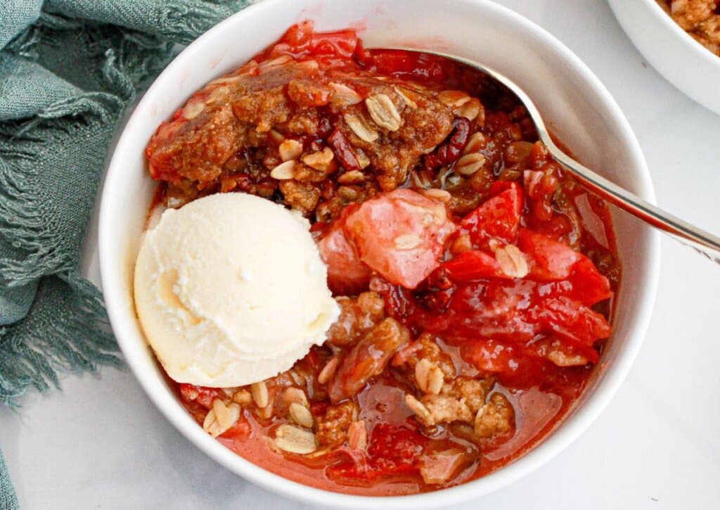 Serving of strawberry rhubarb crisp in a white bowl topped with a scoop of ice cream.