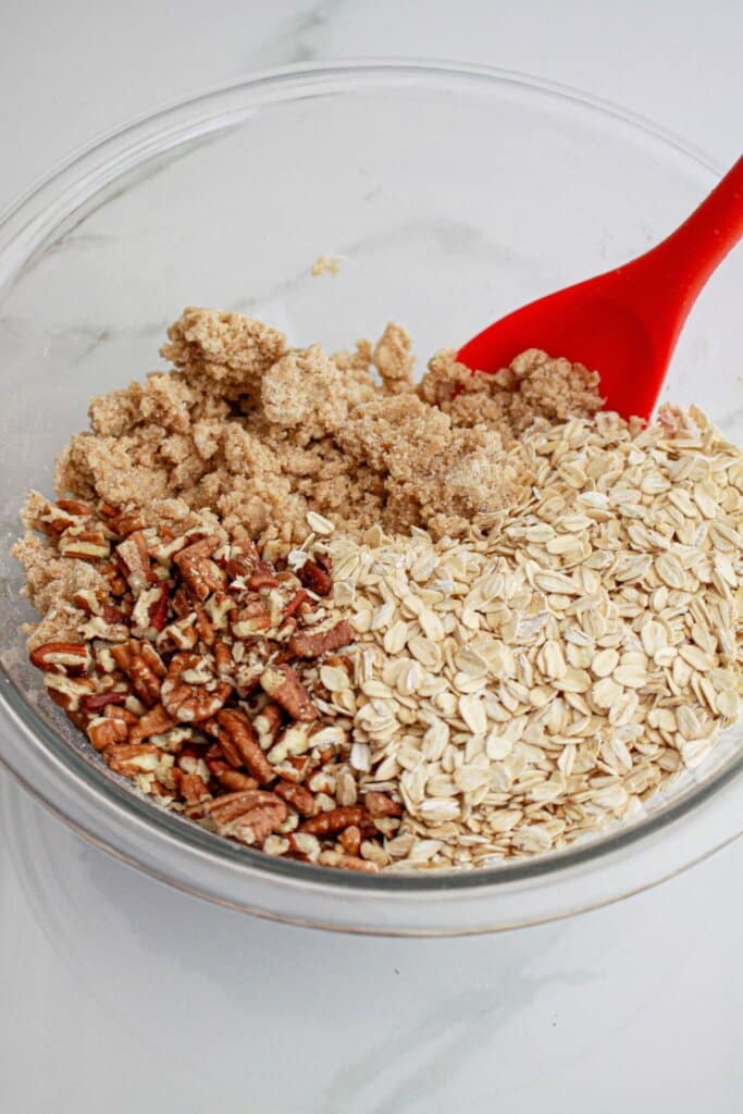 Brown sugar, oats, flour, and pecans being mixed in a glass bowl.