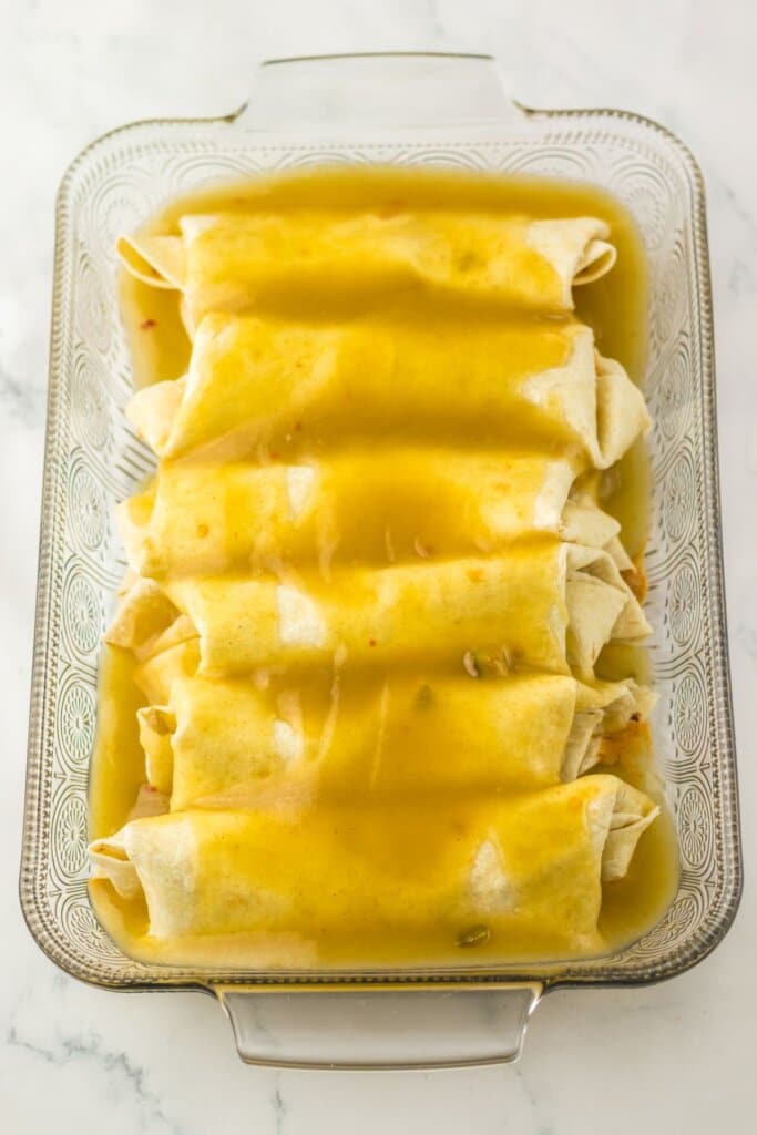 Sauce and cheese on top of rolled tortillas in a baking dish.