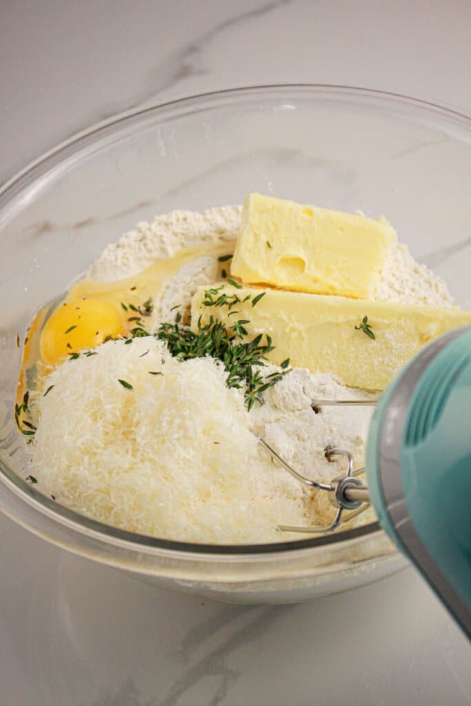 Butter, cheese, egg, flour, and herbs being mixed in a large bowl.