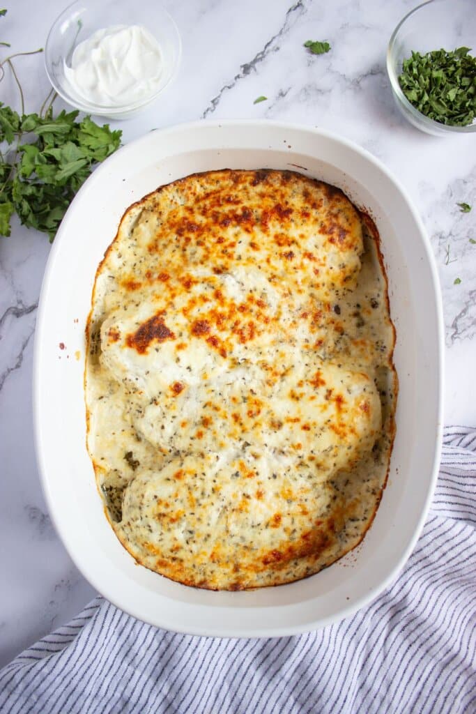 A white baking dish filled with baked cheesy sour cream chicken, golden brown on top with specks of herbs.