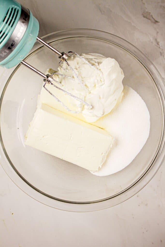 Cream cheese, sour cream, and sugar being mixed into a glass bowl.