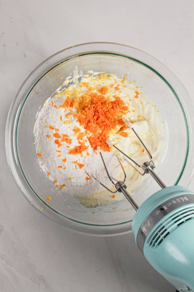Orange and lemon zest being mixed with cream cheese and pudding mix in a glass bowl.