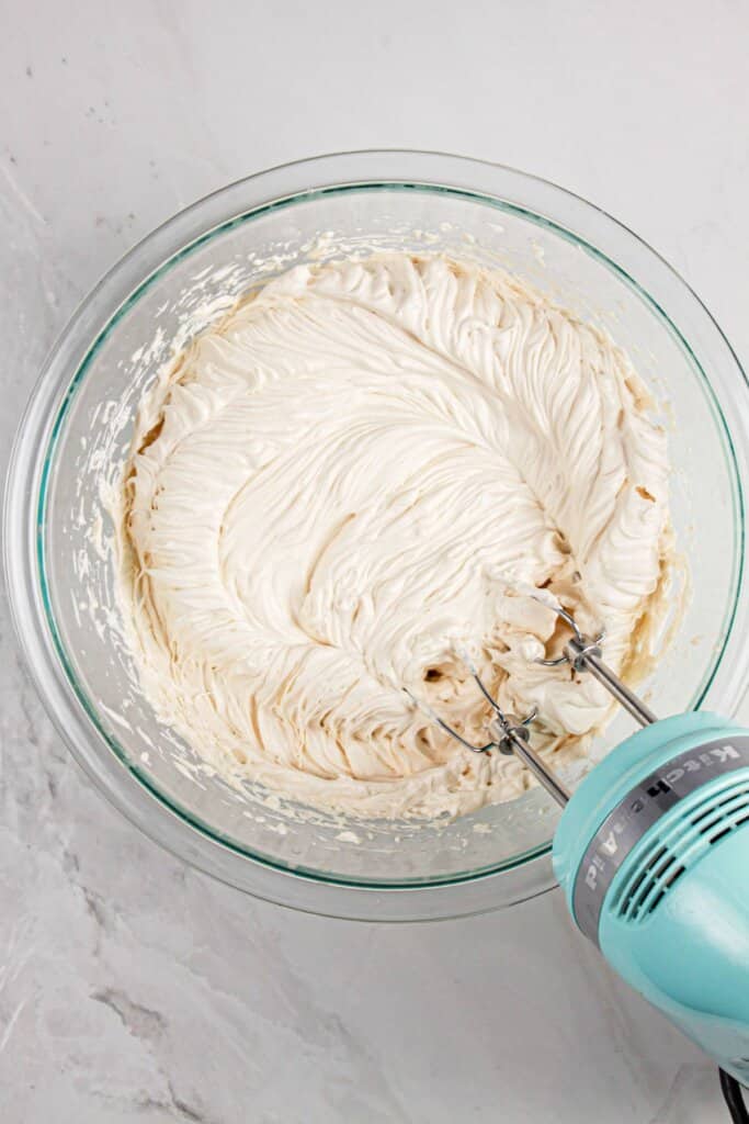 Whipped cream being mixed with a mixer in a glass bowl.