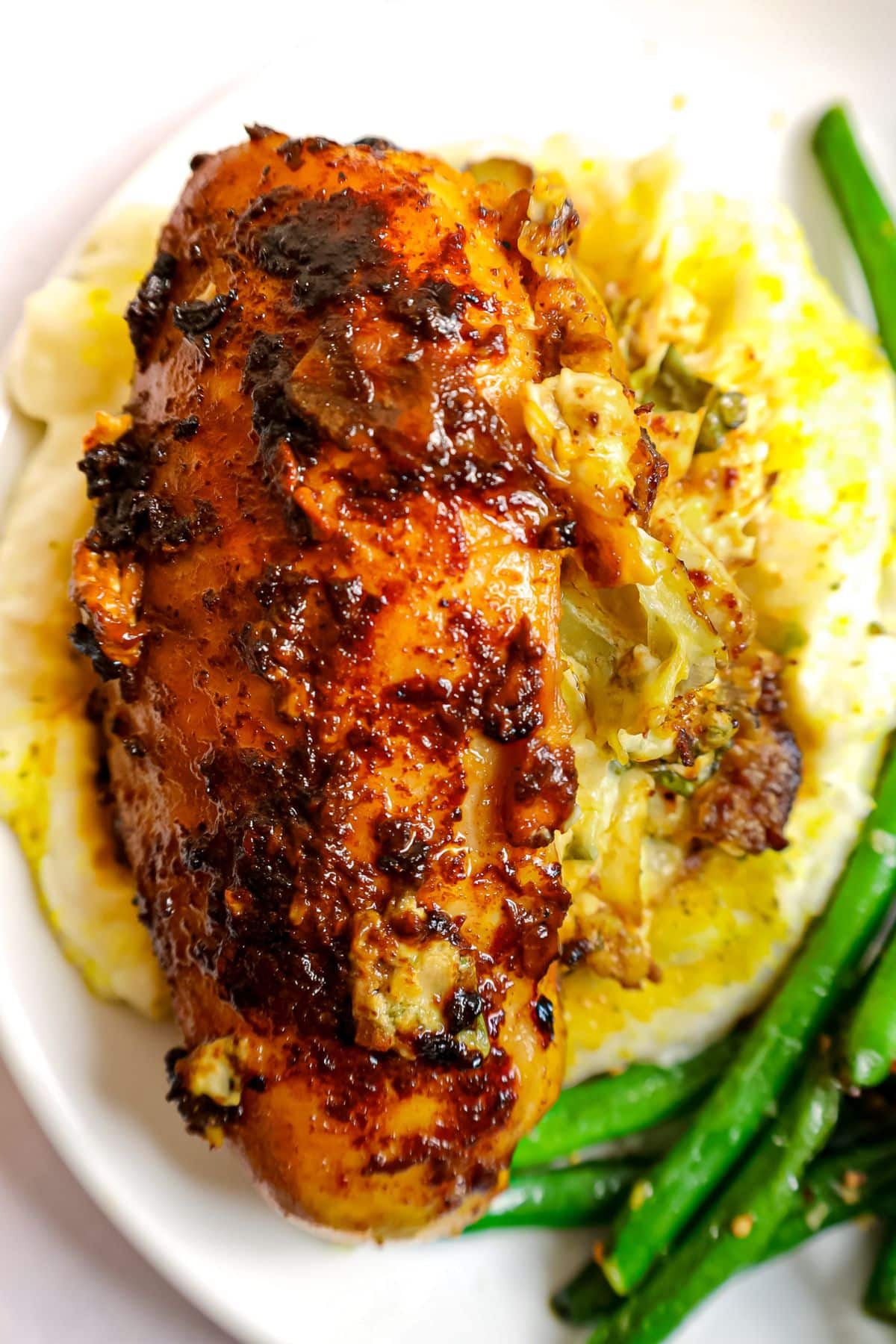 A cooked, seasoned chicken breast served over mashed potatoes with a side of green beans on a white plate.