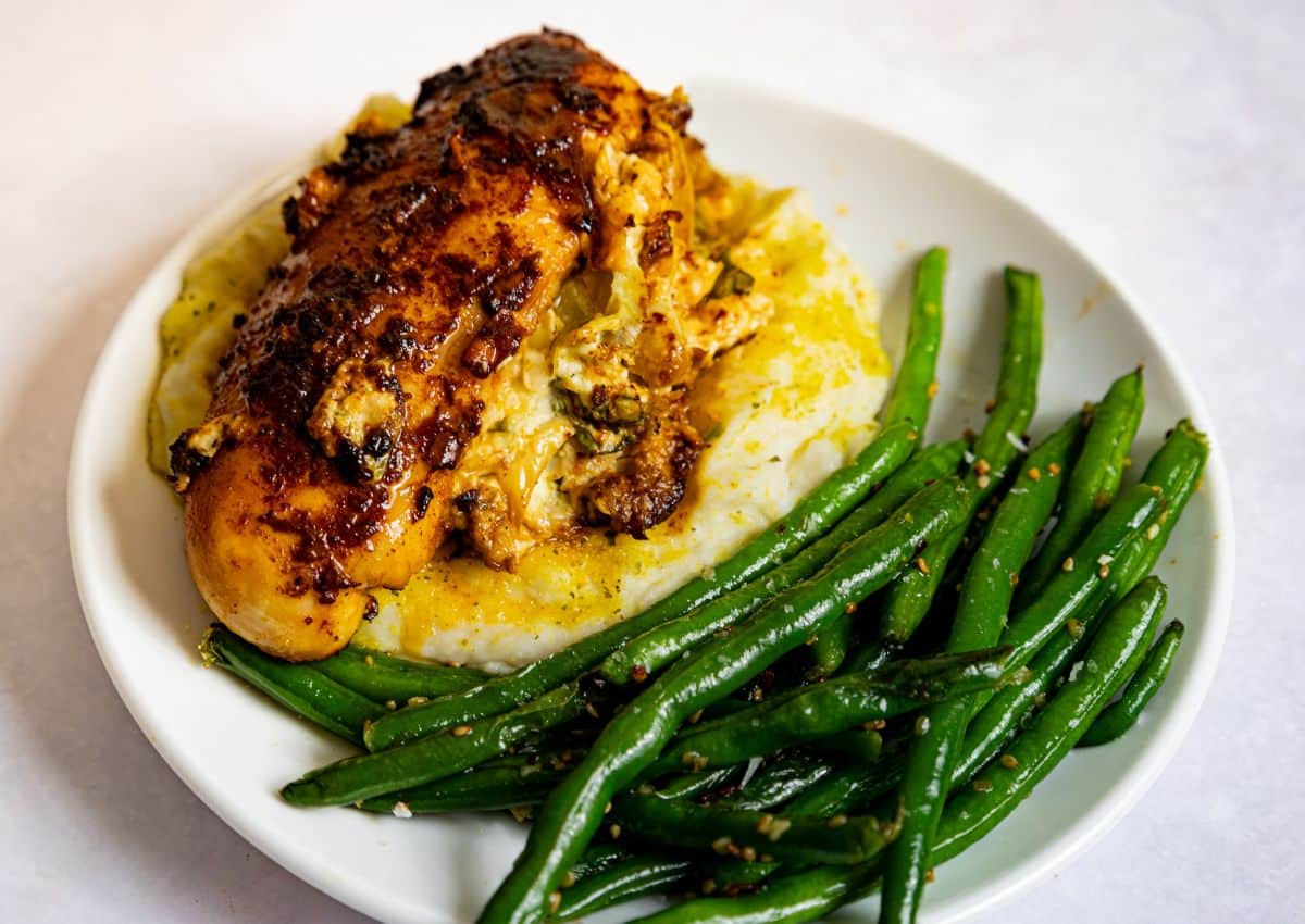 A plate of stuffed chicken breast on mashed potatoes with a side of seasoned green beans.