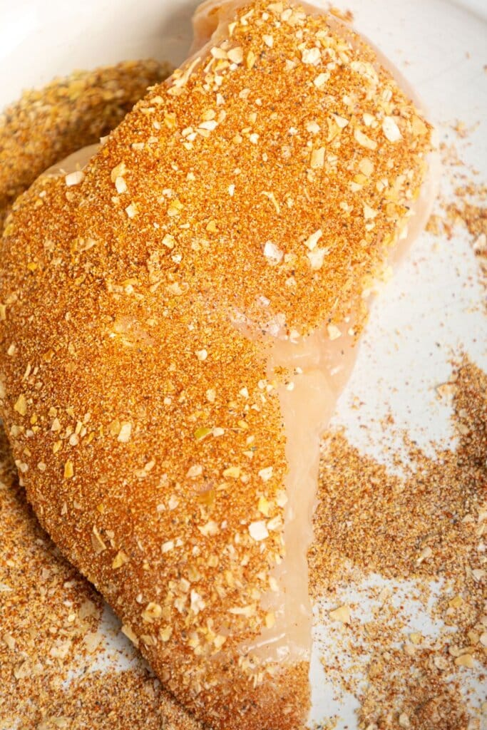 Raw chicken breast coated with a blend of dry seasonings and spices on a white surface.