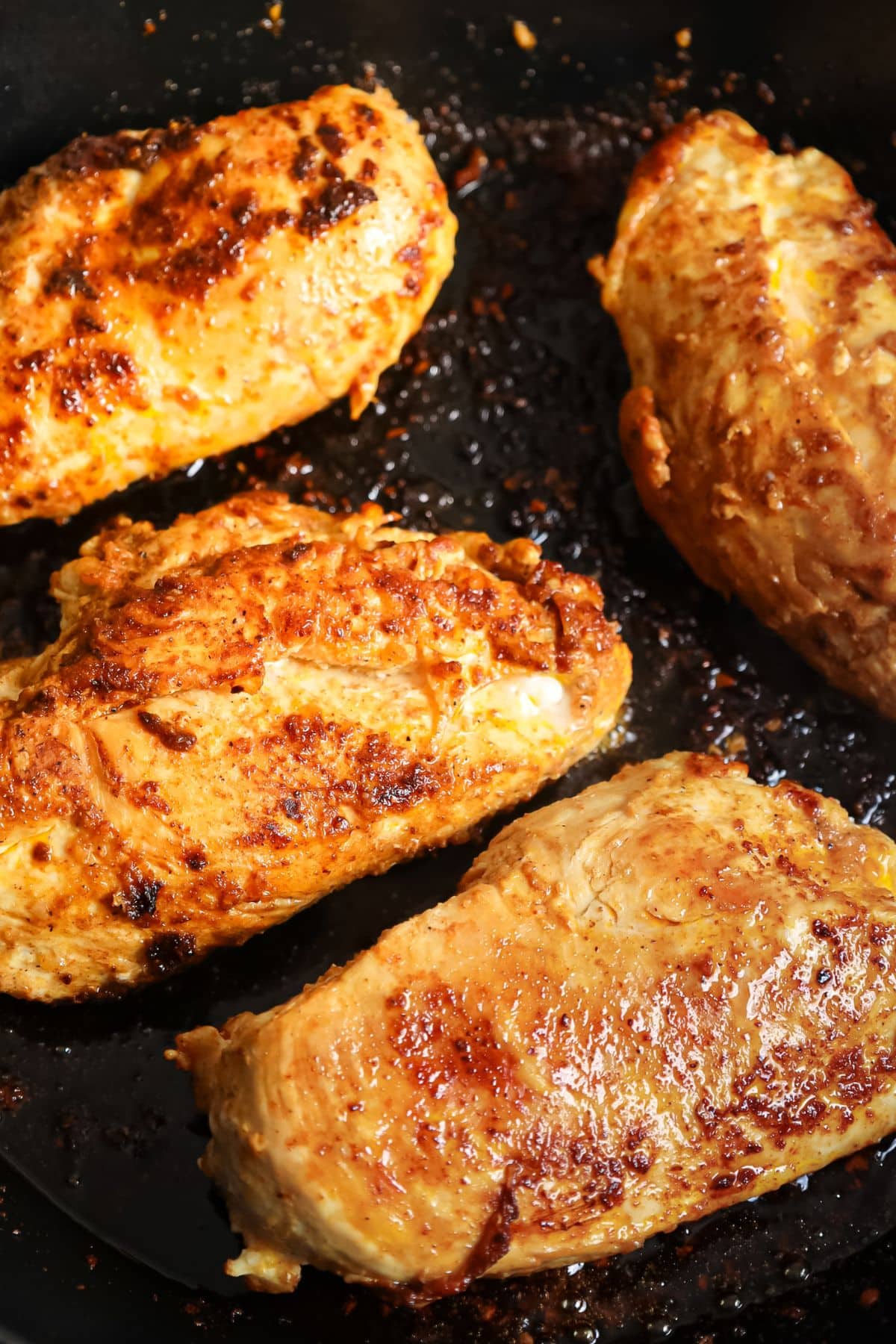 Four browned chicken breasts cooking in a black skillet, showing a seared, seasoned surface.