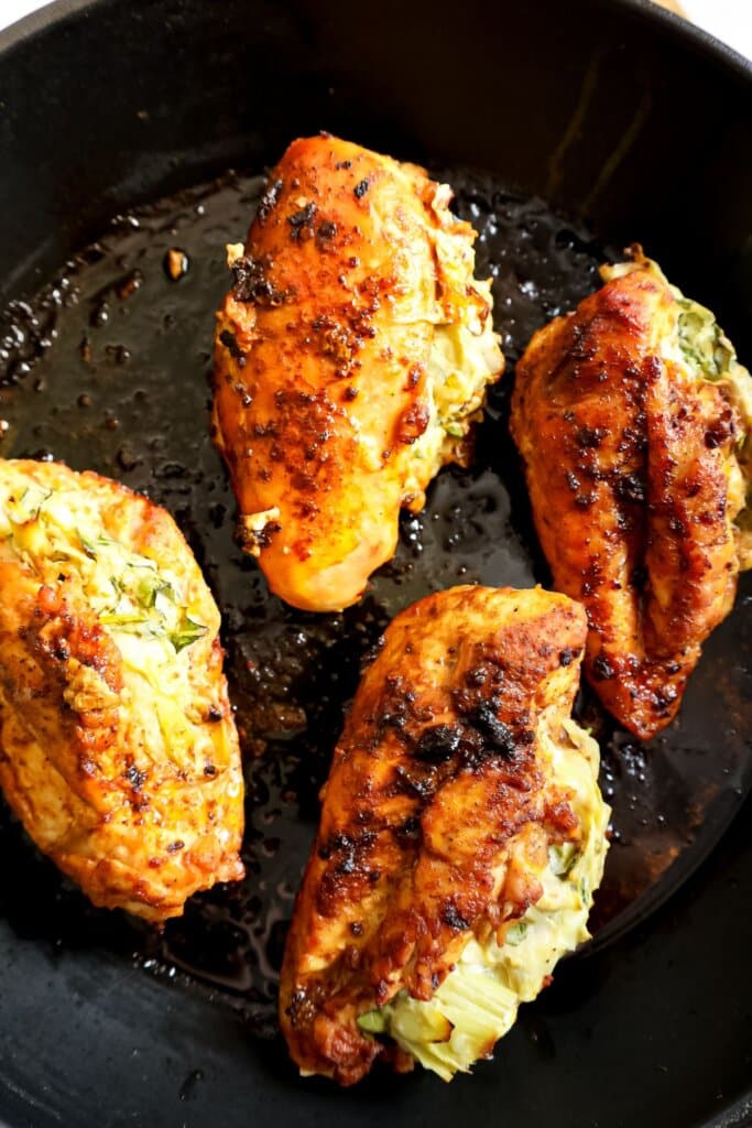 Four seasoned, browned chicken breasts stuffed with vegetables cooking in a black skillet.